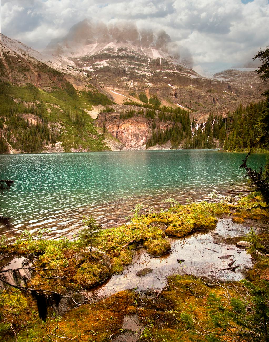 lake at the foot of a steep mountain with a peak in the clouds